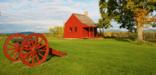 Scenic rural landscape with green lawn, cloud-flecked blue sky, and autumn foliage punctuated by a small, deep red farmhouse and red cannon carriages.